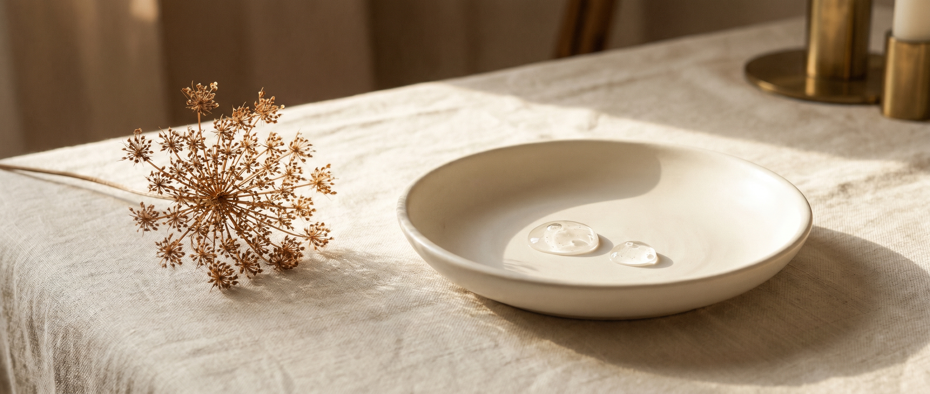 A small porcelain dish holding two glistening crystal-clear gel droplets beside a delicate dried botanical on cream linen — Aesura Health's hyaluronic-acid filler practice (Restylane, Juvederm) in Hackensack, Bergen County NJ.