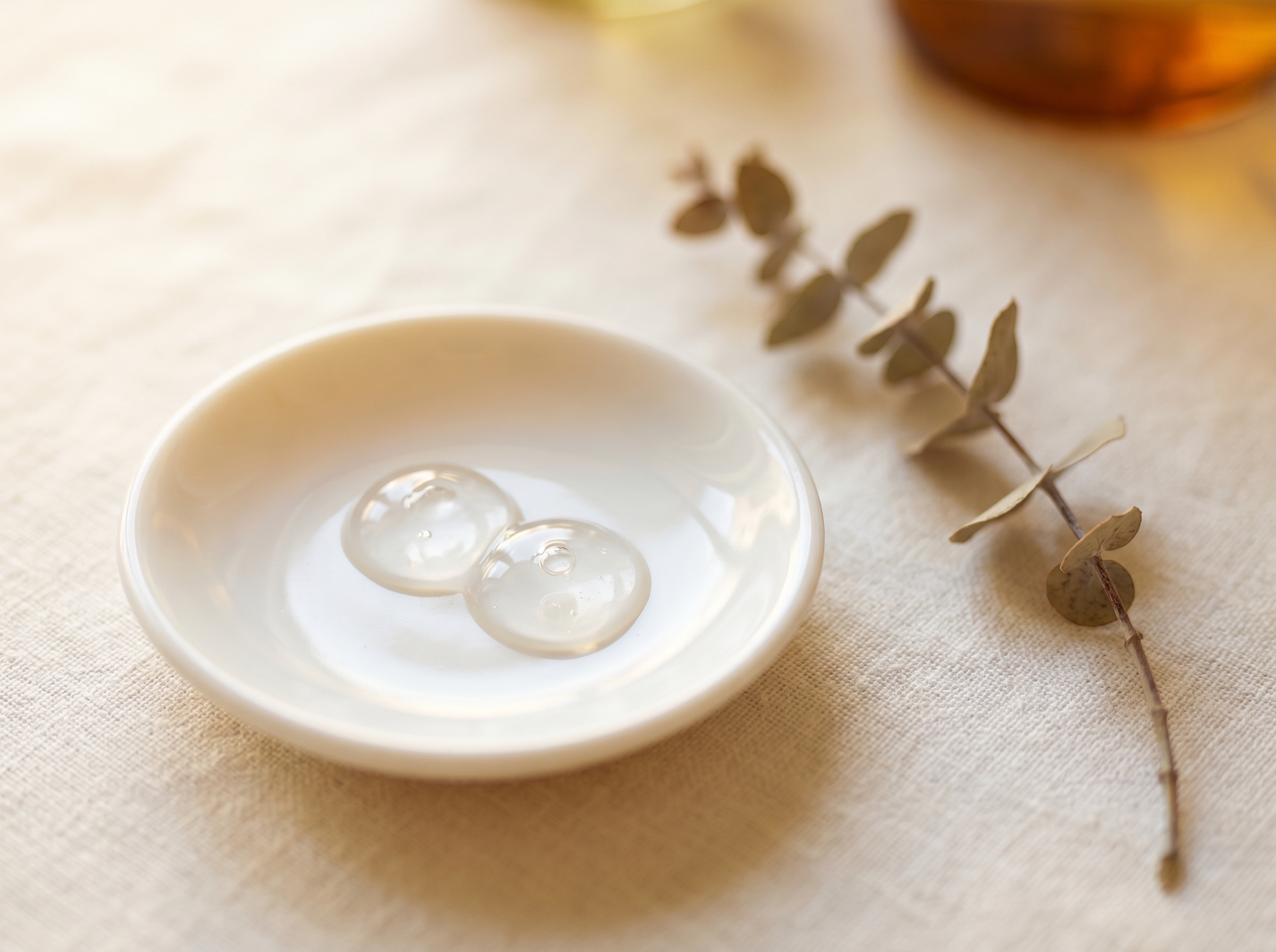 A small porcelain dish holding two clear gel droplets beside a dried botanical stem on cream linen — dermal fillers at Aesura Health